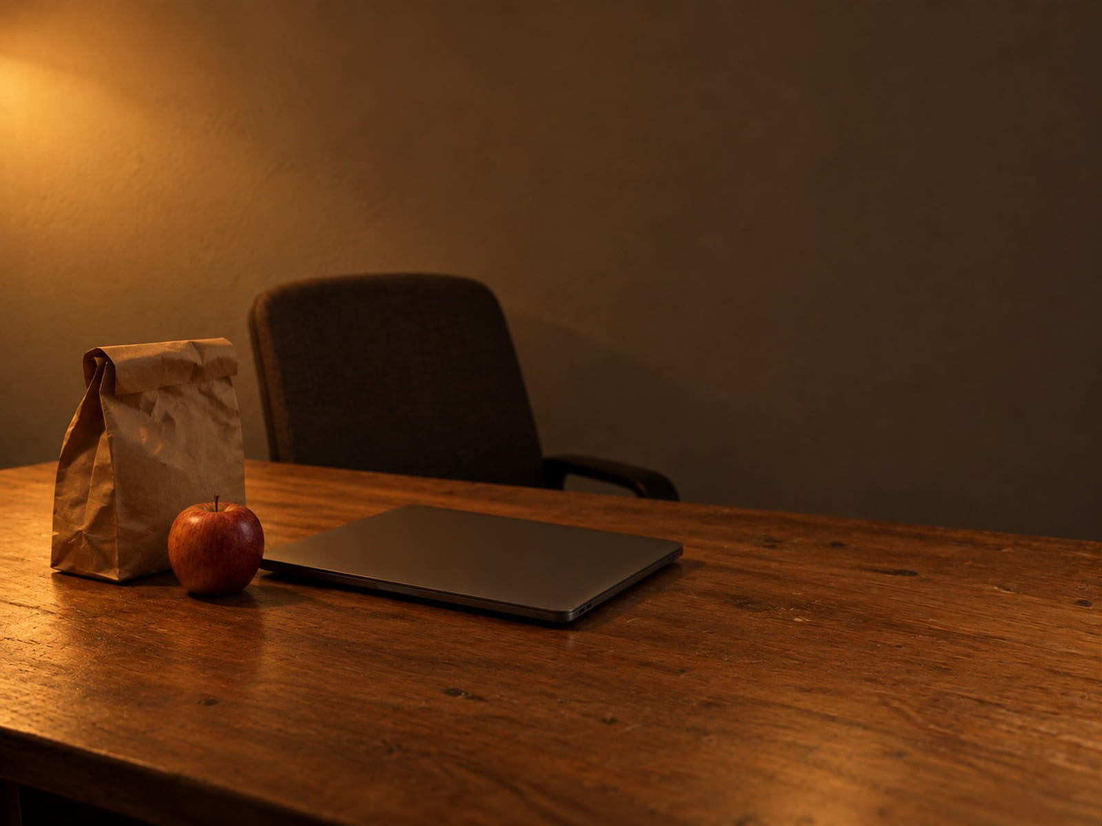 A paper lunch bag and an apple on an office desk beside a closed laptop, the chair pushed back