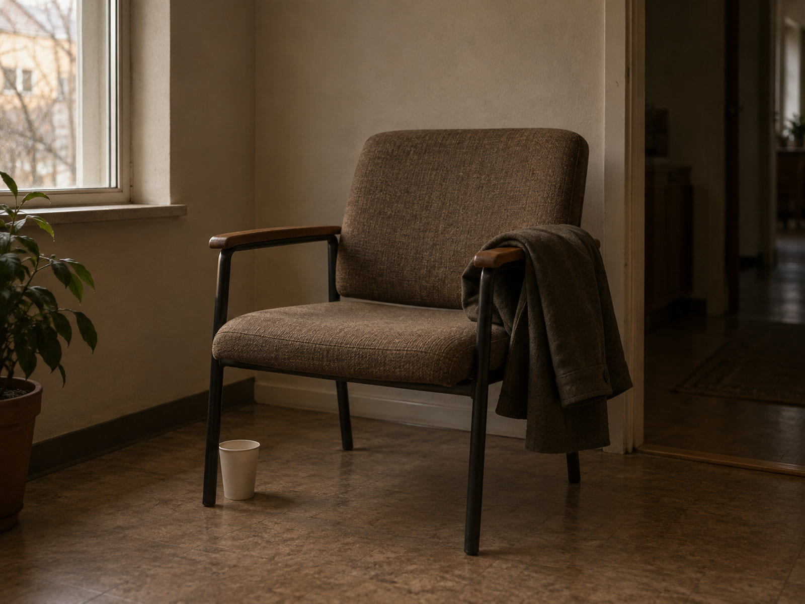 An empty hospital waiting room chair with a folded coat over its arm and a paper cup on the floor beside it