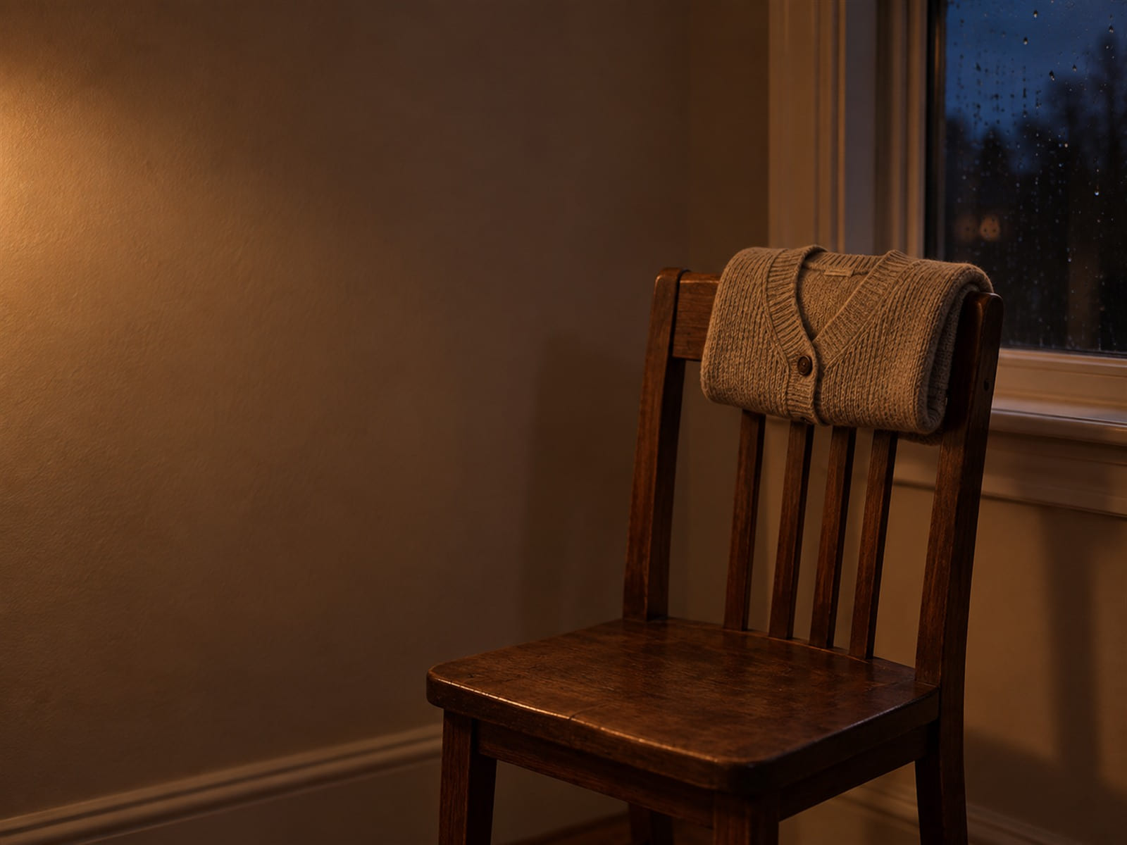 A folded cardigan on the back of an empty chair beside a rain-streaked window