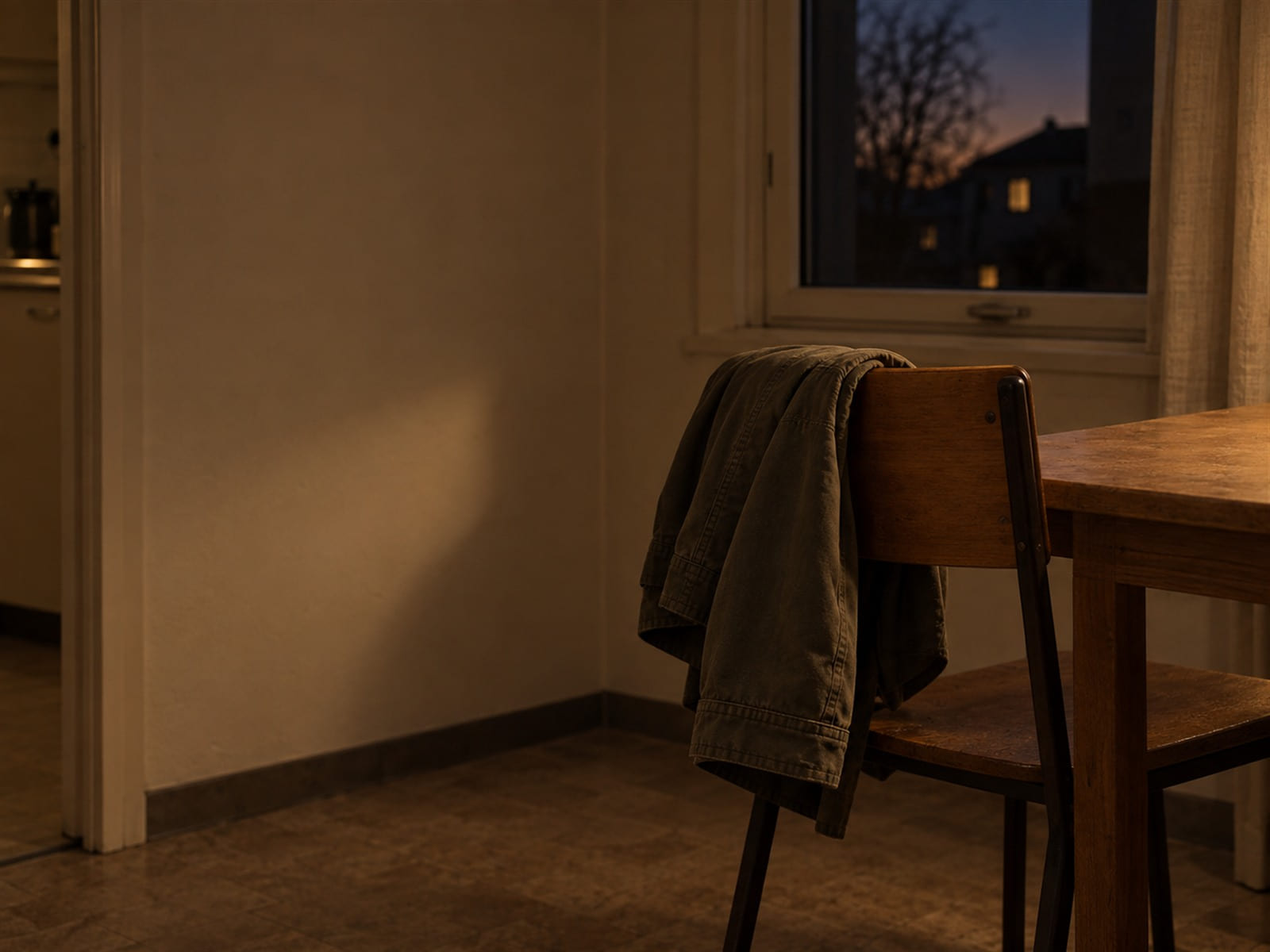 A folded jacket hanging on the back of a desk chair next to a window at dusk