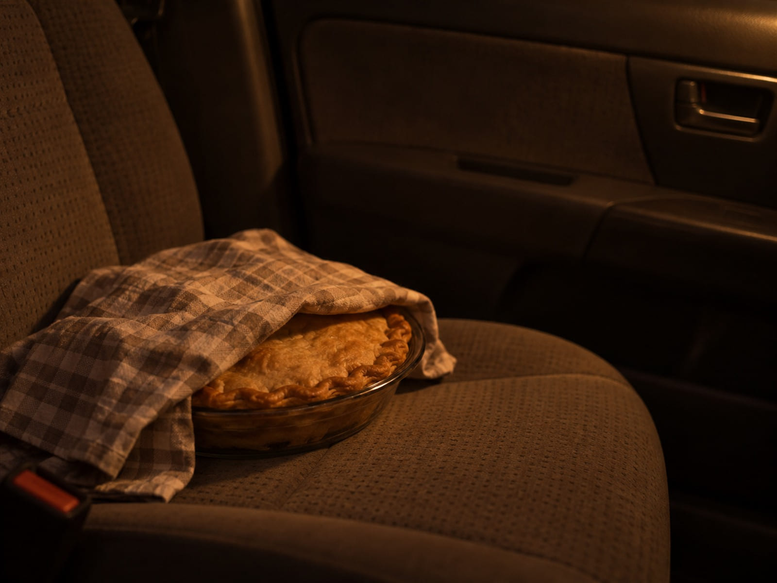 A pie in a glass dish covered with a checked cloth on the passenger seat of a car